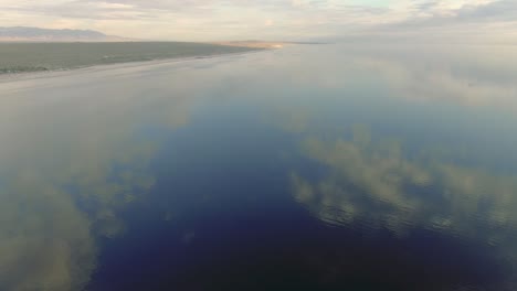 aerial-overview-of-Salton-Sea,-Salt-lake-and-mountains,-salt-flats-reflecting-sun,-clouds-and-blue-sky,-symmetry-reflective-aerial-landscape-panoramic-movement,-view-at-water-reflection-of-the-sky