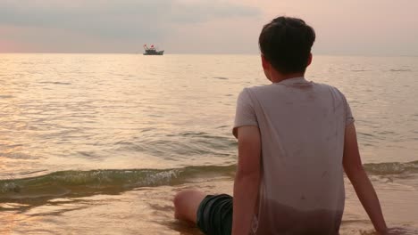 Lonely-serene-young-man-sitting-on-seashore-watching-a-ship-sailing-in-beautiful-sunset