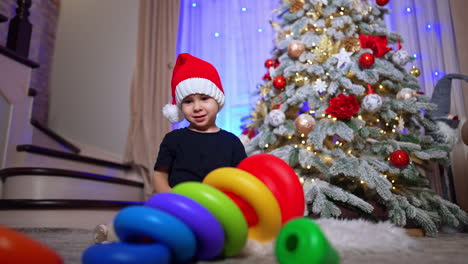 adorable caucasian toddler in red cap sitting near the christmas tree. kid pushes the toy pyramid falling on the floor. low angle view.