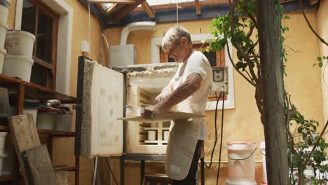 senior caucasian man wearing apron taking fired pots from kiln at pottery workshop