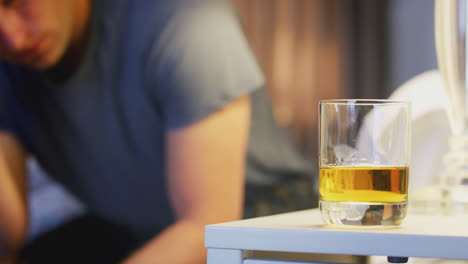 depressed man wearing pajamas sitting on side of bed with glass of whisky on bedside cabinet