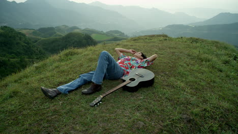 Músico-Masculino-Con-Guitarra-Acústica-Se-Acuesta-En-El-Paisaje-Verde-De-Las-Colinas-Montañosas-Mirando-El-Cielo,-Usando-Ropa-Colorida-Vintage