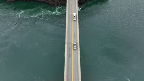 Top-down-aerial-of-cars-passing-over-a-bridge-with-vibrant-blue-water-underneath