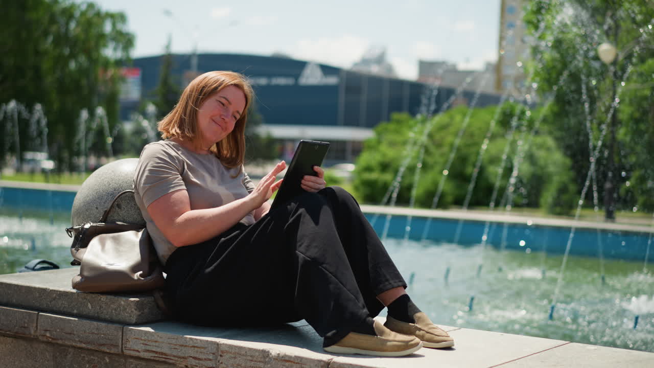Smiling woman sitting relaxed by city fountain using tablet under bright summer sunlight, legs folded, bag by her side, surrounded by sparkling water and greenery