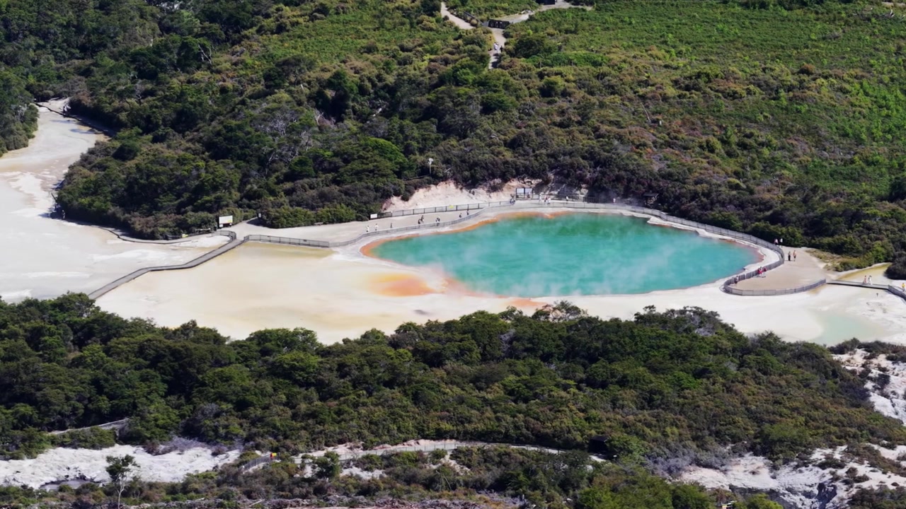 Aerial view of a geothermal hot spring in Rotorua, Bay of Plenty, New Zealand, surrounded by native forest and colorful mineral terraces, showcasing volcanic landscapes and geothermal activity.