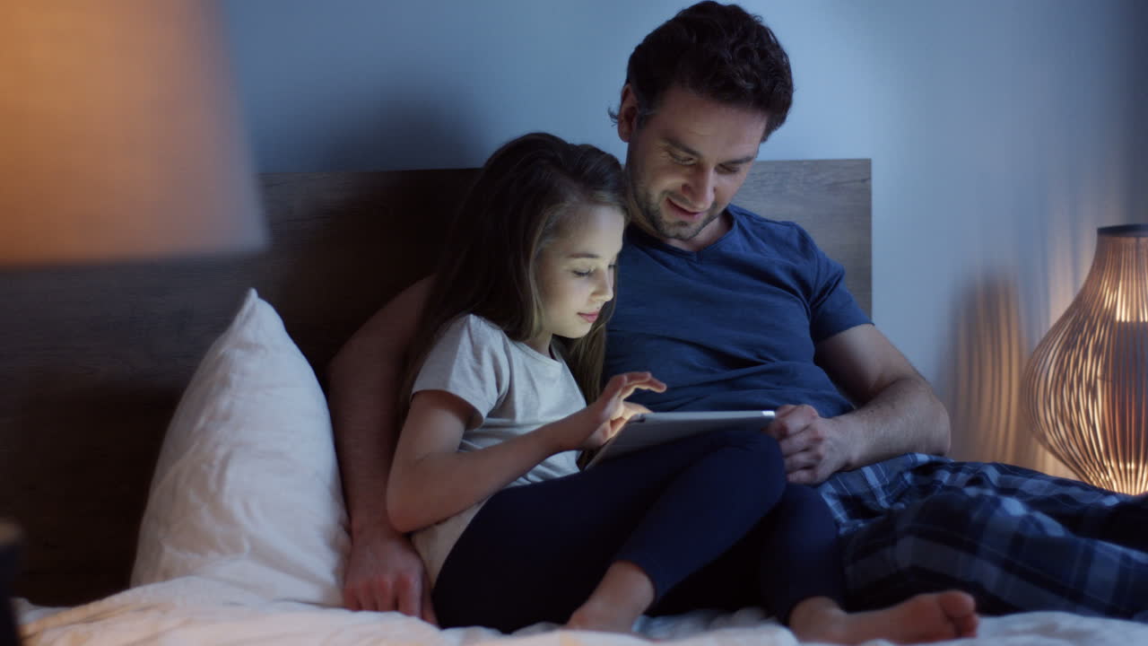 Caucasian father lying on the bed with his cute little daughter scrolling and taping on the tablet at night