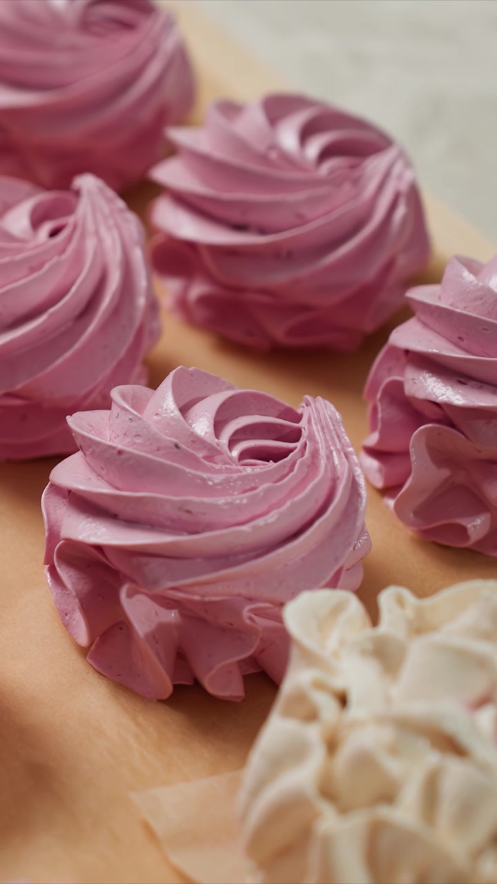 Primer plano de galletas de merengue rosas y blancas dispuestas. Las galletas muestran superficies lisas y brillantes con bayas visibles. Superficie de madera debajo de los productos horneados con formas de flores dispersas.