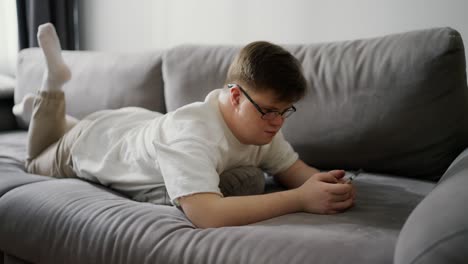 Un-Joven-Con-Síndrome-De-Down-Usando-Un-Teléfono-Celular-Mientras-Descansa-En-El-Sofá-De-Casa