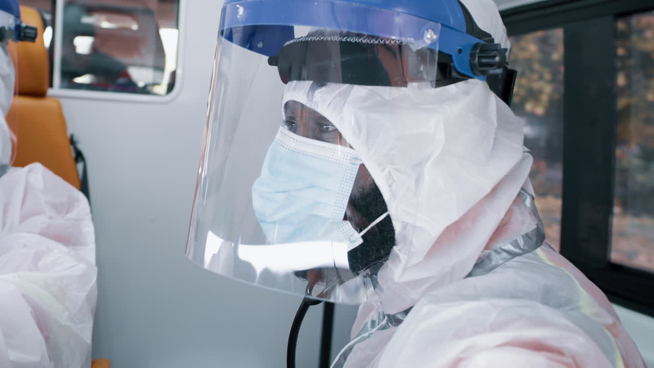 black Paramedic Using Tablet in Ambulance during Pandemic African American Doctor in Hazmat Suit Browsing Tablet while Sitting Near Window of Ambulance on Ride to Hospital during Epidemic