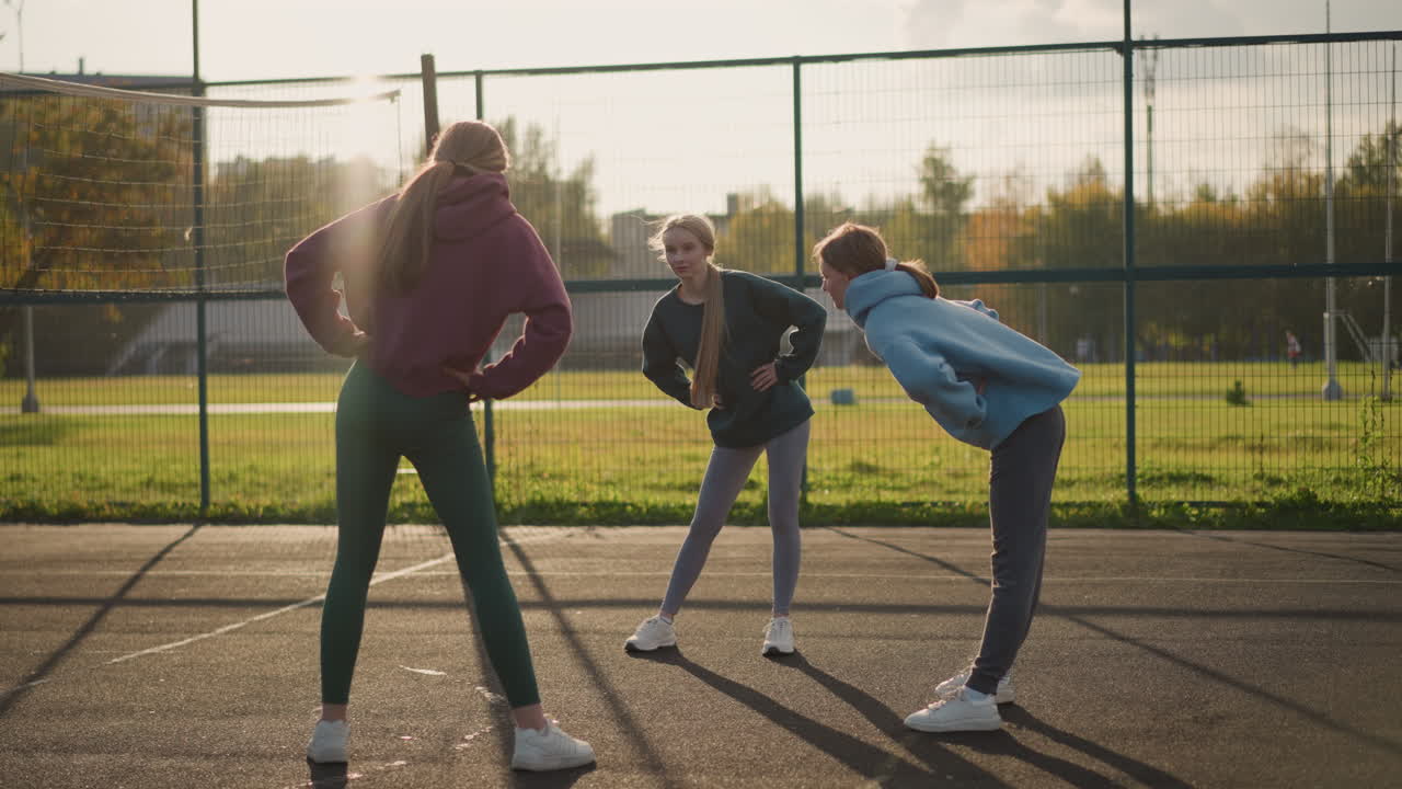 레과 후디를 입은 여자들이 야외 코트에서 피트니스 활동을 하며, 주거 건물과 축구장을 배경으로 신체 운동과 팀워크를 강조합니다.