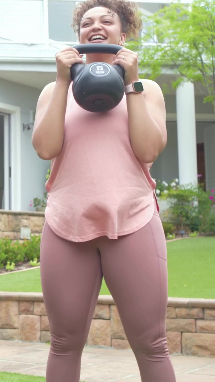 Video verticale: Donna con top rosa che solleva un kettlebell da 5 kg e fa goblet squat su un patio per il fitness.