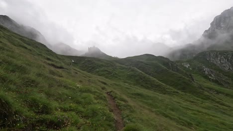 a winding alpine trail leads through mist-covered green slopes toward a silhouetted fortress structure shrouded in fog, creating a mysterious and dramatic mountain atmosphere in the italian alps