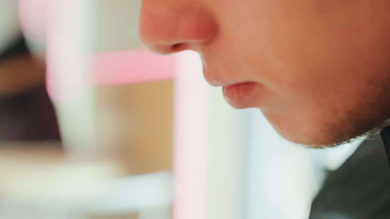 Nærbillede profil af ung mand, der spiser stille i blødt neonlys på café, tætte skud af læber, hage og skægstubbe, langsomme mundbevægelser, delikate detaljer af gaffel og ske, varm pastel bokeh, eftertænksom stemning.