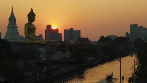 Breathtaking-sunset-behind-a-giant-golden-Buddha-statue-next-to-a-tranquil-river-as-a-boat-sails-by