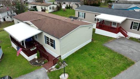 Small-mobile-home-in-trailer-park,-bungalow,-manufactured-home-close-to-other-houses,-patriotic-American-flag,-beautiful-landscaping,-United-States-of-America-USA-aerial-establishing-shot-in-winter