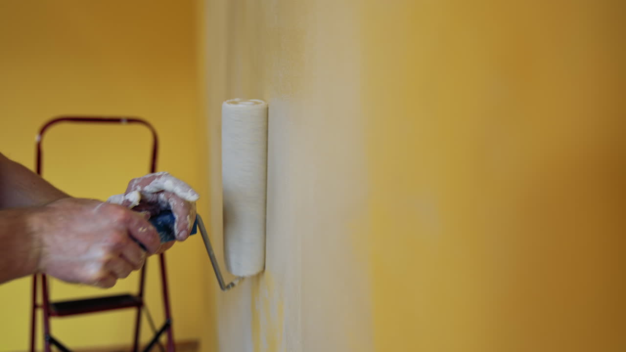 Hand with paint roller during room renovation. Man moving paint roller with white color on the wall. Professional painting services. Close-up.
