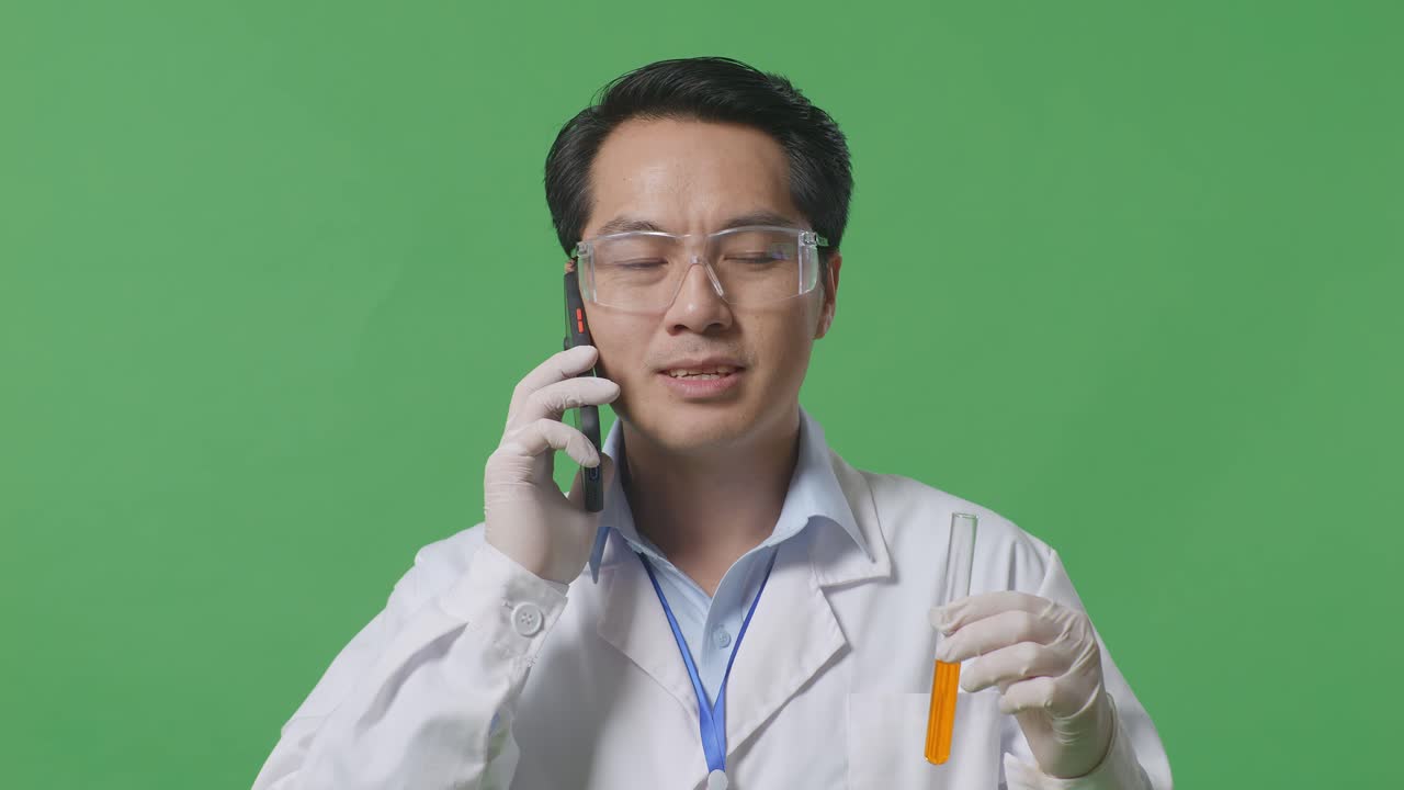 연구실의 녹색 스크린 배경에 서서 스마트폰에 대화하는 동안 그의 손에 실험 튜브에 오렌지 액체를보고있는 아시아 남성 과학자의 근접