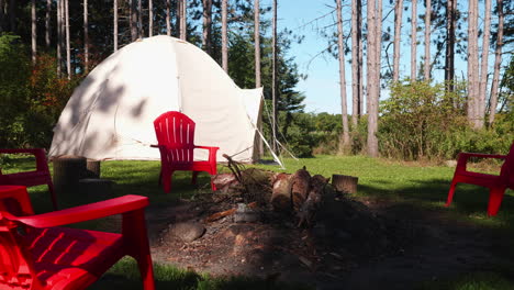 A-few-red-plastic-chairs-are-standing-near-a-bonfire-in-the-forest,-where-a-tent-is-placed-in-the-background