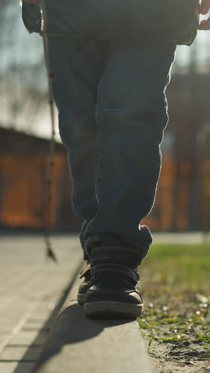 un primer plano de un niño caminando por una acera sosteniendo un palo, perdió su paso antes de subir de nuevo a la acera el niño está vestido con una chaqueta negra brillante, rodeado de
