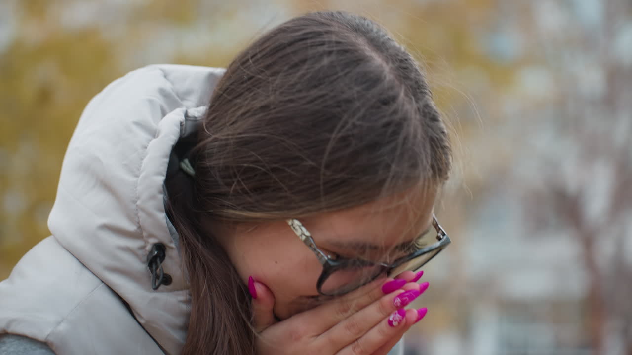 Close up of young woman wearing glasses with long hair swaying in wind smiling gently and nodding in excitement with polished nails visible and blurred golden autumn trees creating warm background