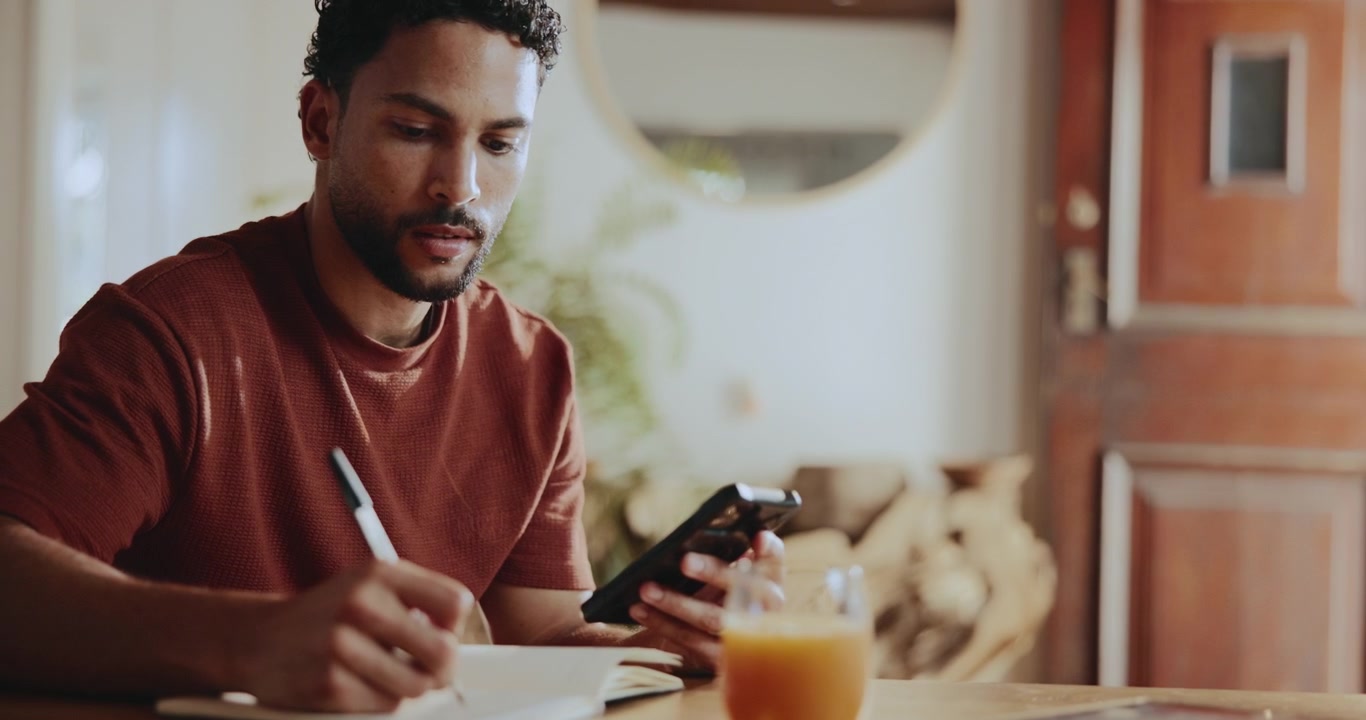 Giovane uomo che usa lo smartphone e scrive su un quaderno a casa