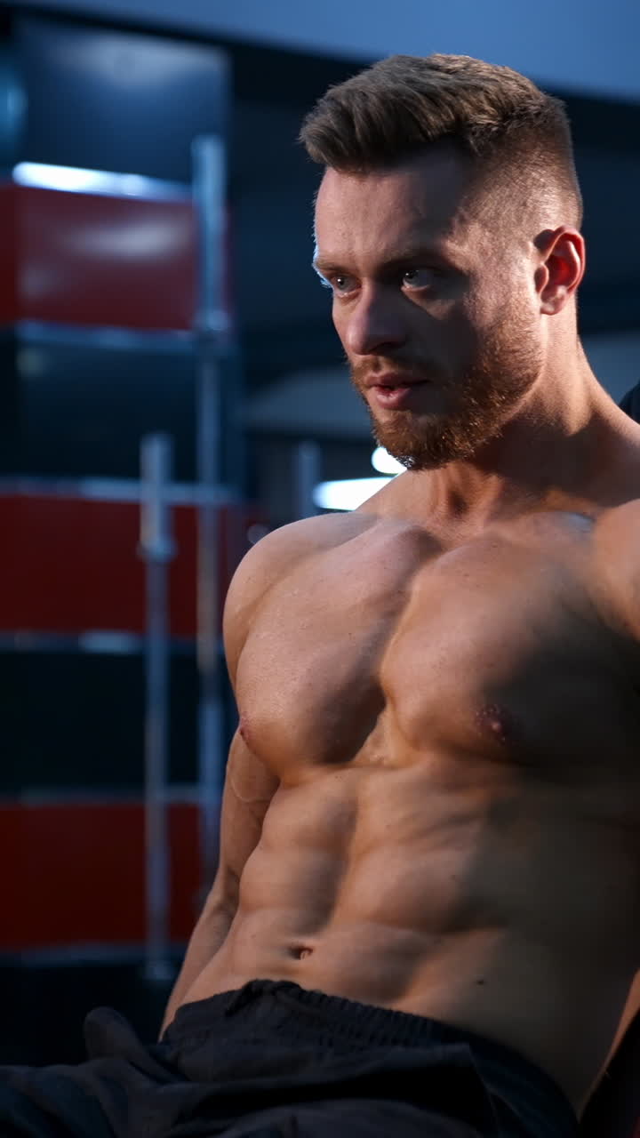 Giovane bodybuilder a torso nudo si allena con i manubri. Addominali scolpiti. Filtro luce blu. Video di lato. Video verticale.