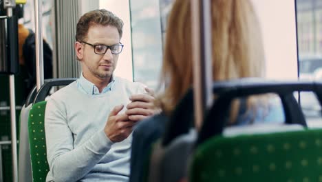 Joven-Apuesto-Con-Anteojos-Tocando-Y-Enviando-Mensajes-De-Texto-En-El-Teléfono-Inteligente-Mientras-Está-Sentado-En-El-Tranvía-De-Camino-A-Casa-O-Al-Trabajo