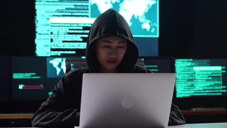 asian female hacker in a hood looking at camera while works on a laptop with maps and data on display screens of multiple monitors in a dark office room