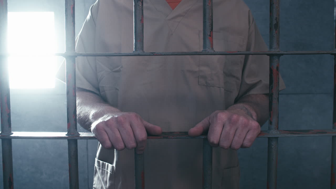 средний кадр тюремной решетки: заключенный подходит к двери своей камеры, держится за решетку и уходит с экрана