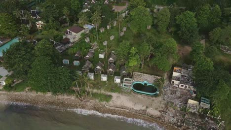Aerial-birds-eye-view-reveal-shot-panning-out-from-a-abandoned-and-derelict-beach-bungalow-tourist-resort-to-the-mountains-on-Koh-Chang-in-Thailand-with-effects-of-covid-on-global-travel-and-tourism