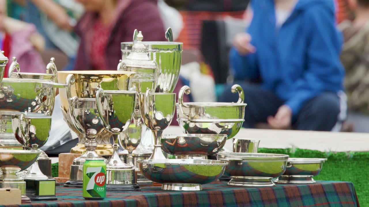Multiple polished silver trophies and awards are arranged on a tartan-covered table outdoors, with spectators in the background. Daylight, shallow depth of field, slow camera pan