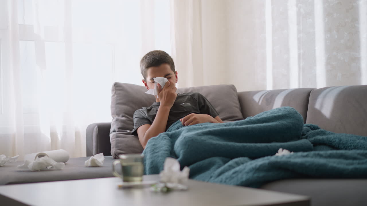 Young boy sneezing into tissue with tissue on table and couch, tissues littered all over, and glass cup on table, sick child resting under blanket at home, managing cold symptoms