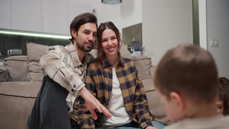 Happy-brunette-man-with-stubble-in-a-checkered-beige-shirt-hugs-his-brunette-wife-in-a-white-t-shirt-and-checkered-shirt-while-they-watch-their-little-children-being-creative-and-drawing-after-moving-to-a-new-apartment-in-a-modern-apartment