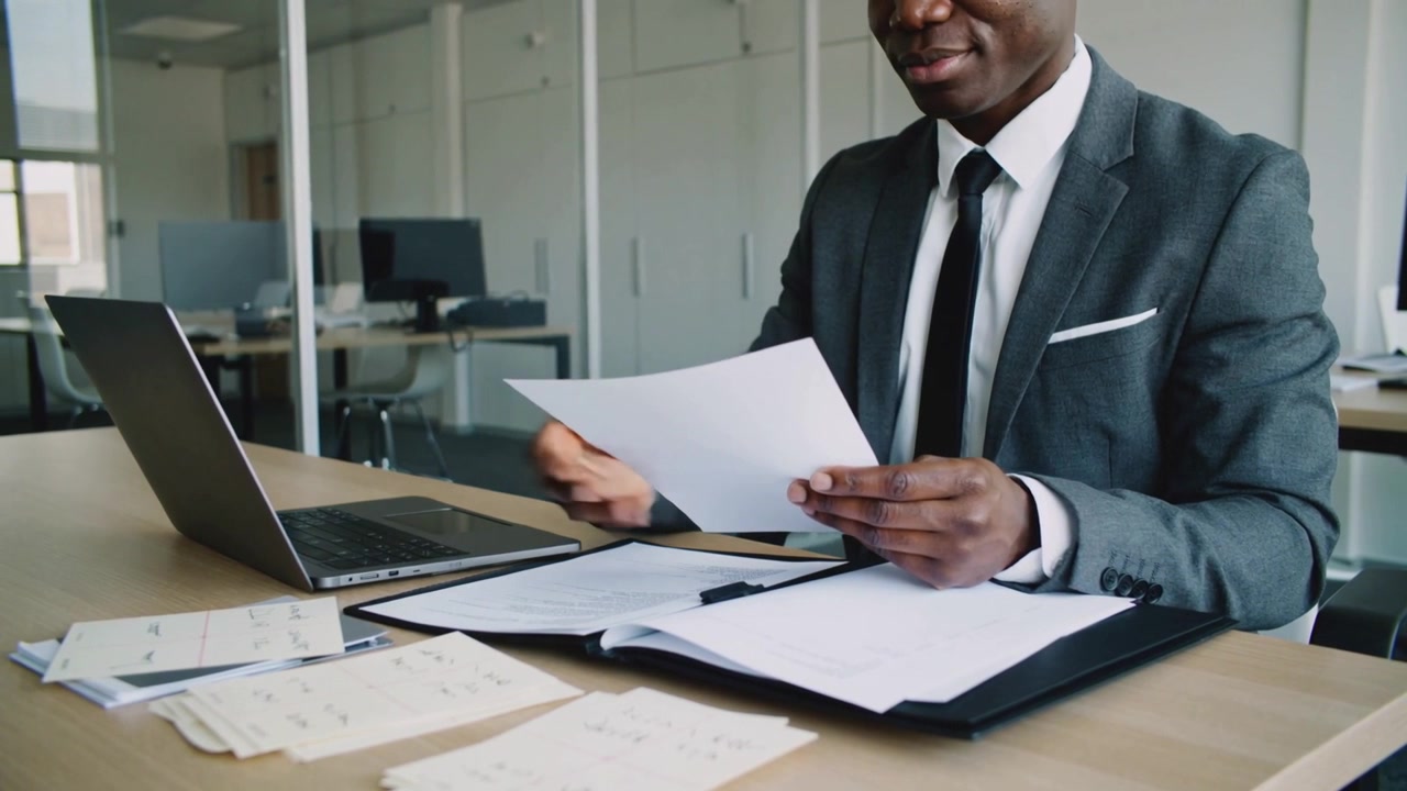 Reviewing manager in gray suit lifting sheet, sorting papers at desk with laptop folder, copy space