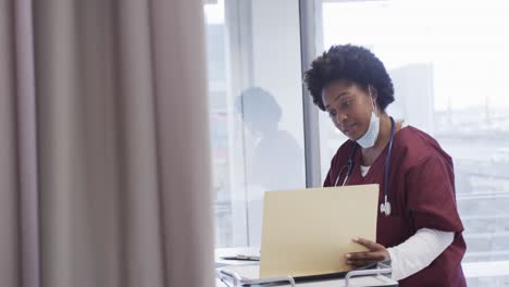 African-american-female-doctor-with-face-mask-and-taking-notes-in-hospital-room,-slow-motion