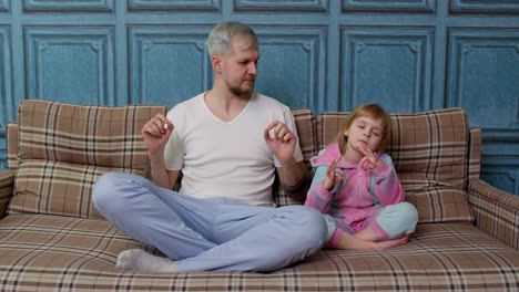 father and little daughter kid are meditating while sitting in yoga lotus pose on sofa at home room