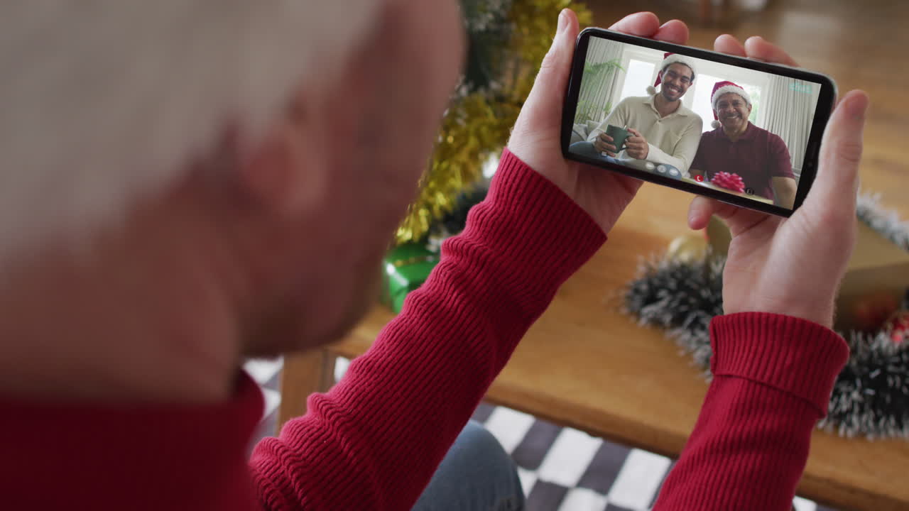 hombre caucásico con sombrero de santa usando un teléfono inteligente para una videollamada de navidad con una familia sonriente en la pantalla