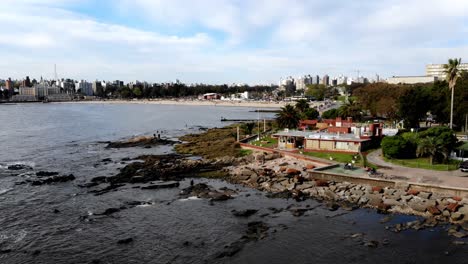 Aerial-view-of-montevideo-uruguay-pier-with-the-city-in-the-background,-rocks-and-waves-crashing-on-a-sunny-day