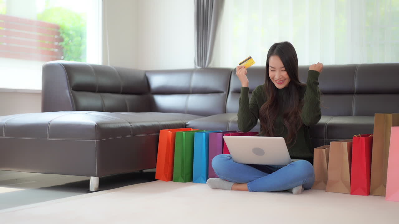 Asian female model sitting on floor with laptop and credit card surrounded by colorful shopping bag cheer and smiles while ordering another gift. Lady shopping only excited for a deal and discount
