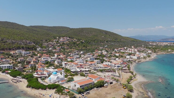 Filmato con drone che cattura il villaggio di Skala sull'isola di Agistri, con folle vivaci sulla spiaggia sabbiosa, acque turchesi, architettura tradizionale greca, colline lussureggianti e chiara luce estiva