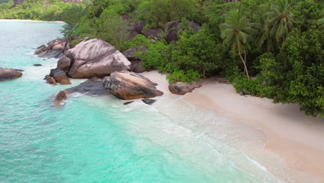 Drone-shot-of-white-sandy-beach,-large-granite-stone,-trees-and-waves-crashing-on-the-shore,-Mahe-Seychelles-60-fps