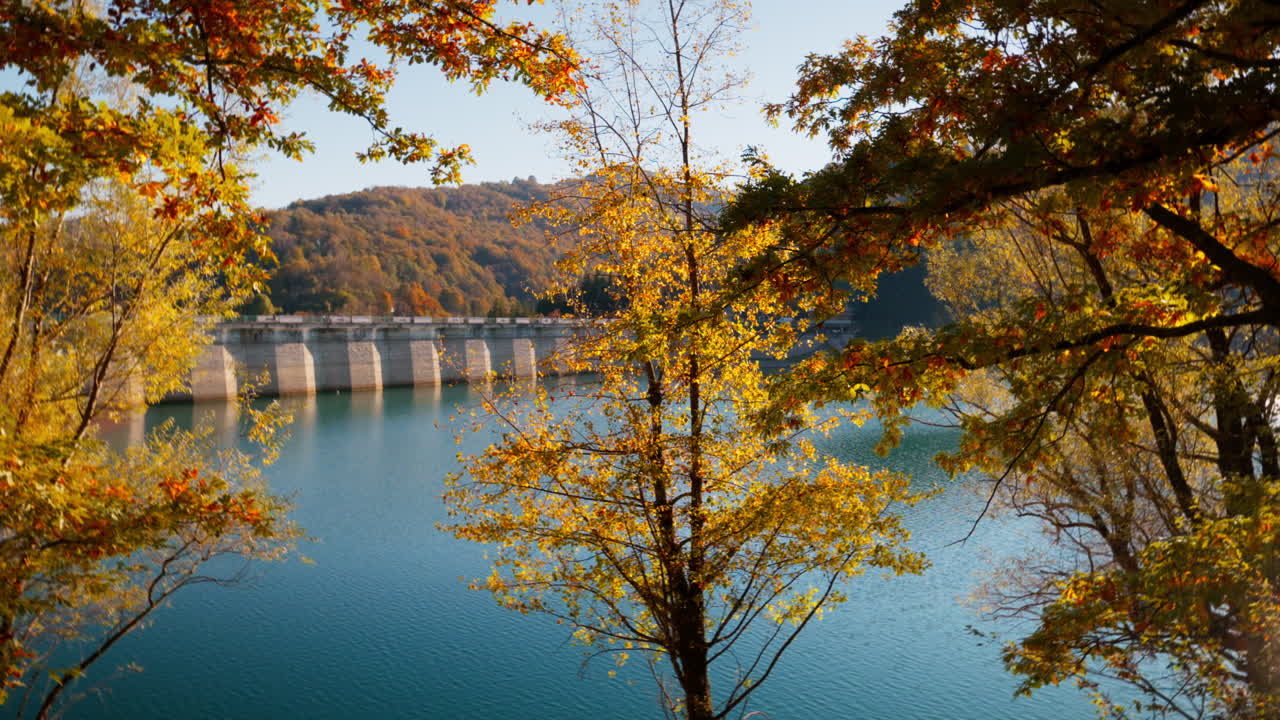 Tranquil autumn lake with colorful trees and distant bridge in sunlight