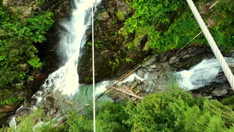 A-POV-shot-of-a-white-men-in-blue-boots,-balancing-over-a-steel-cable-bridge,-across-a-wild-river-and-a-waterfall