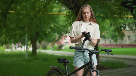 young adult adjusting black glove while standing next to her parked bicycle on paved path, surrounded by lush greenery and blurred view of residential building in background