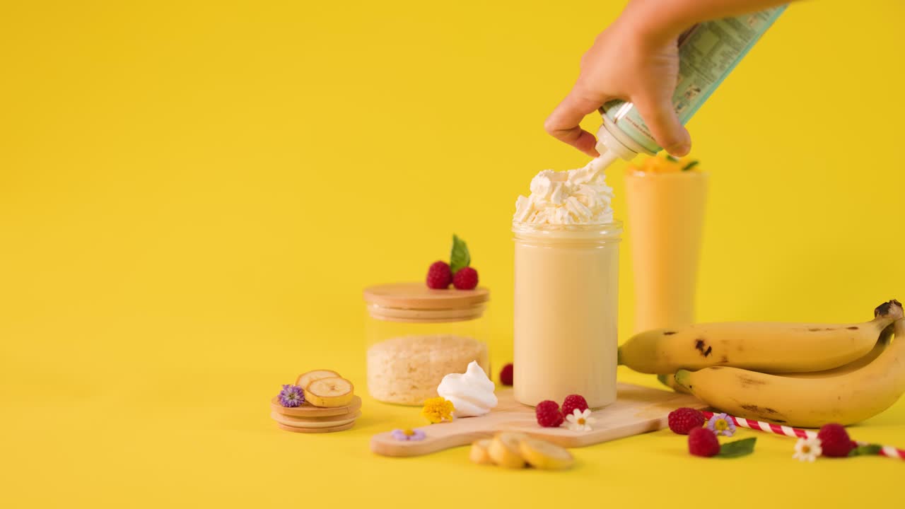 Mani che guarniscono un frullato di banana con panna montata tra frutta tropicale su sfondo giallo brillante
