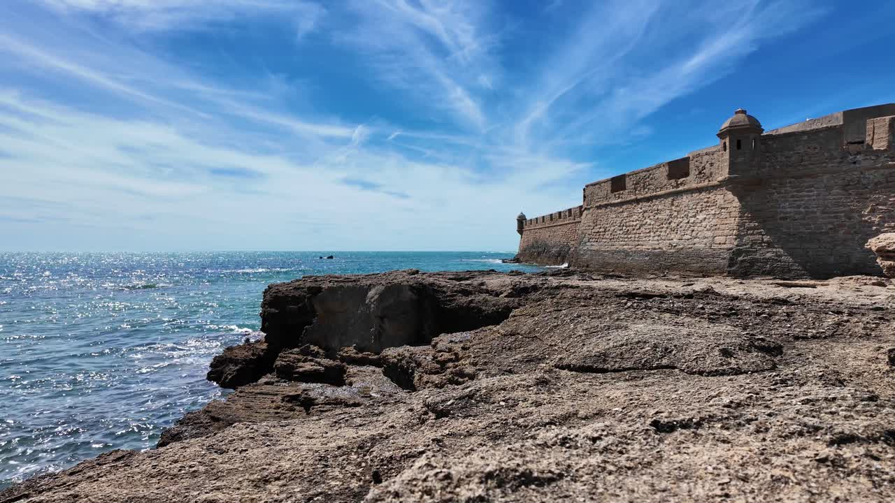 Naturskjønn utsikt over en historisk steinfestning med utsikt over Atlanterhavet i Cádiz, Spania. Bølger slår mot den steinete kystlinjen under en lys blå himmel, noe som fremhever byens maritime arv