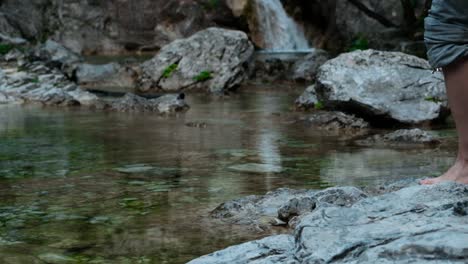 Hiker-With-Barefoot-Crossing-The-Shallow-River-With-Scenic-Waterfall-In-The-Background---close-up-slowmo