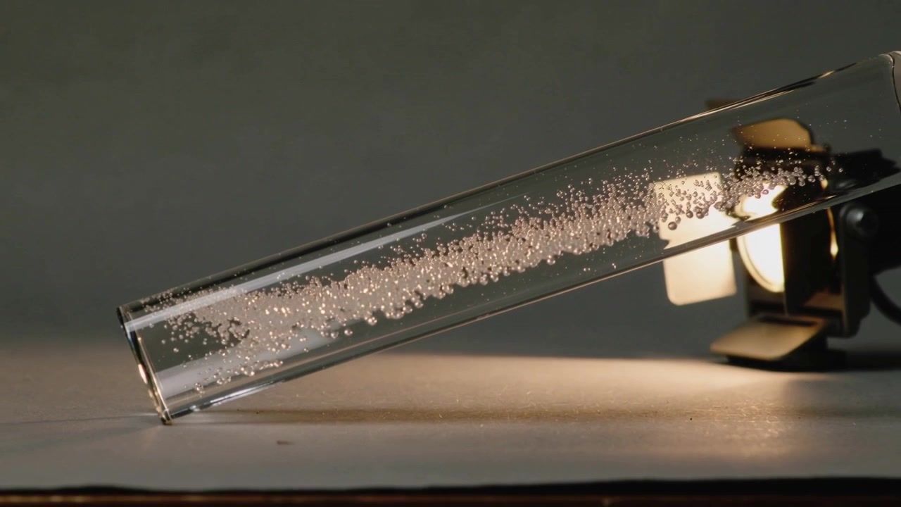 Showing glass tube with clear liquid, bubbles starting, growing and moving toward lit lamp on bench