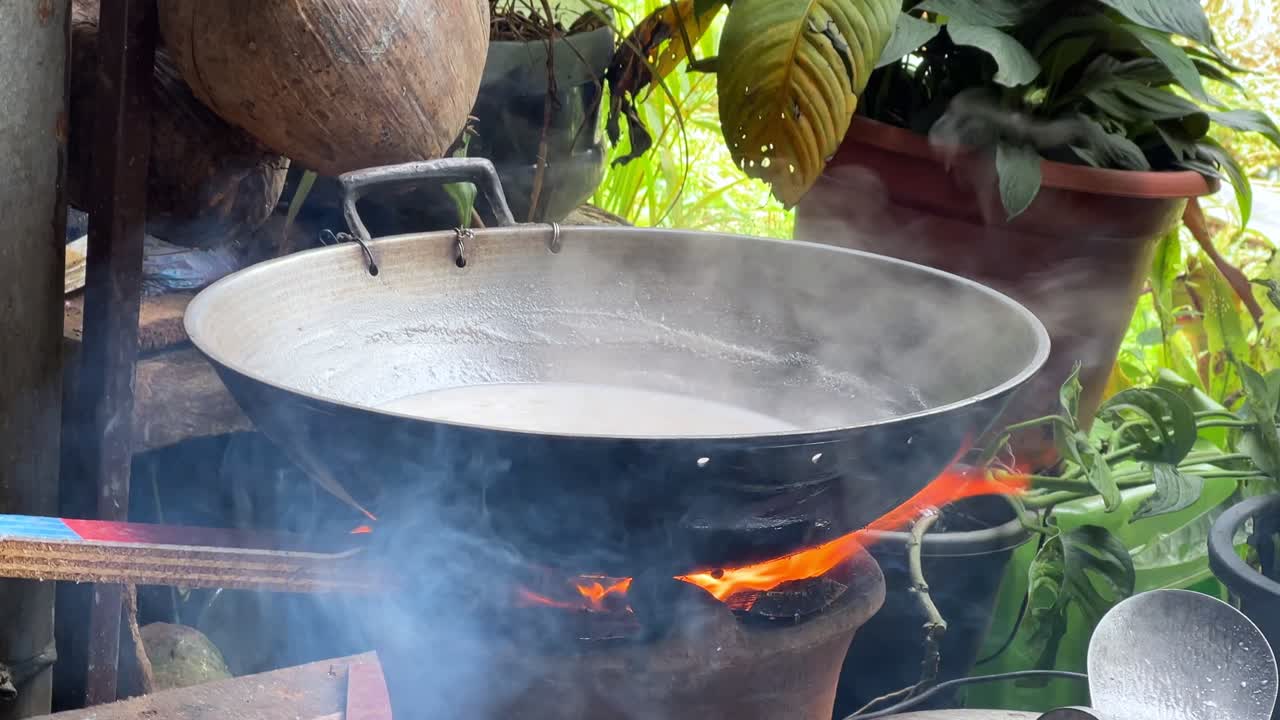 Latte di cocco bolle in una pentola di metallo su un fornello a legna, con denso vapore che sale in una cucina tropicale all'aperto. Ottimo per progetti culinari, culturali o sulla vita rurale