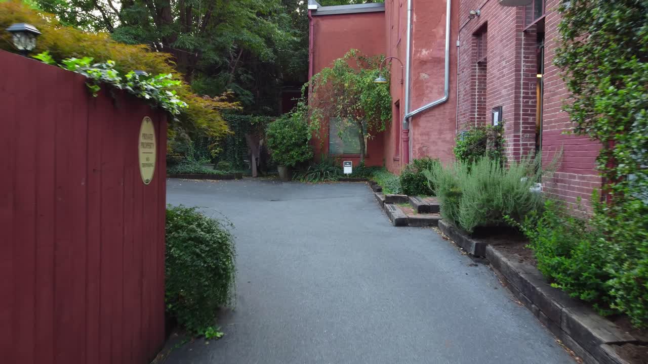 A smooth drone shot glides through a rustic alleyway into a courtyard, framed by red brick on the right, a red fence on the left, and ivy vines weaving across the frame—blending nature and structure.